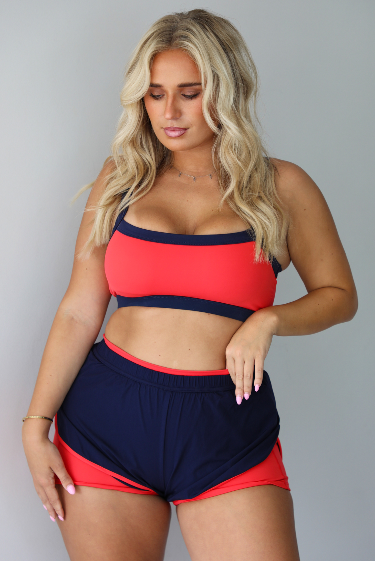 A woman modeling a two-piece athletic set with a bright coral-red and navy blue color-blocked design, standing indoors against a white wall.