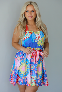 Woman wearing a multi-colored and multi-patterned mini dress with thick red straps, a cinched waistline with a removable tie, and a flowy skirt indoors against a white wall.