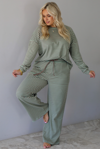 A woman wearing a pale green and brown striped two piece set featuring an oversized long sleeved top and high waisted, wide leg pants, standing indoors against a white wall with white marble flooring. 
