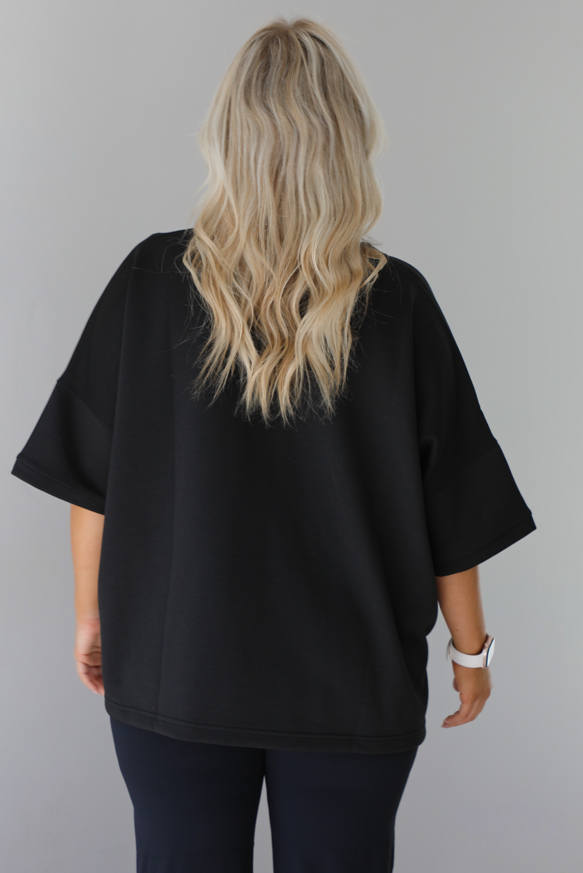 Woman wearing a black oversized box tee with longer short sleeves, a high-low design, and split hem details on either side. The tee is paired with black flared leggings. The photo shows a closeup of the back of the tee. The photo is taken indoors against a white wall.