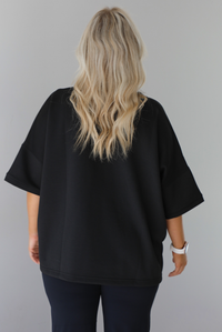 Woman wearing a black oversized box tee with longer short sleeves, a high-low design, and split hem details on either side. The tee is paired with black flared leggings. The photo shows a closeup of the back of the tee. The photo is taken indoors against a white wall.