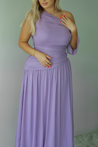 Woman wearing a lavender formal gown featuring a one strap that has extra mesh material that goes to the other arm and drapes down, has a fitted drop waist, and a long, maxi length skirt. The photo is taken indoors against a white wall. The photo shows a closeup of the bodice of the dress. 