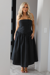 Woman model wearing a strapless/sleeveless, black with white polka dots, maxi length dress indoors. It has a fitted bodice with a flowy skirt. 