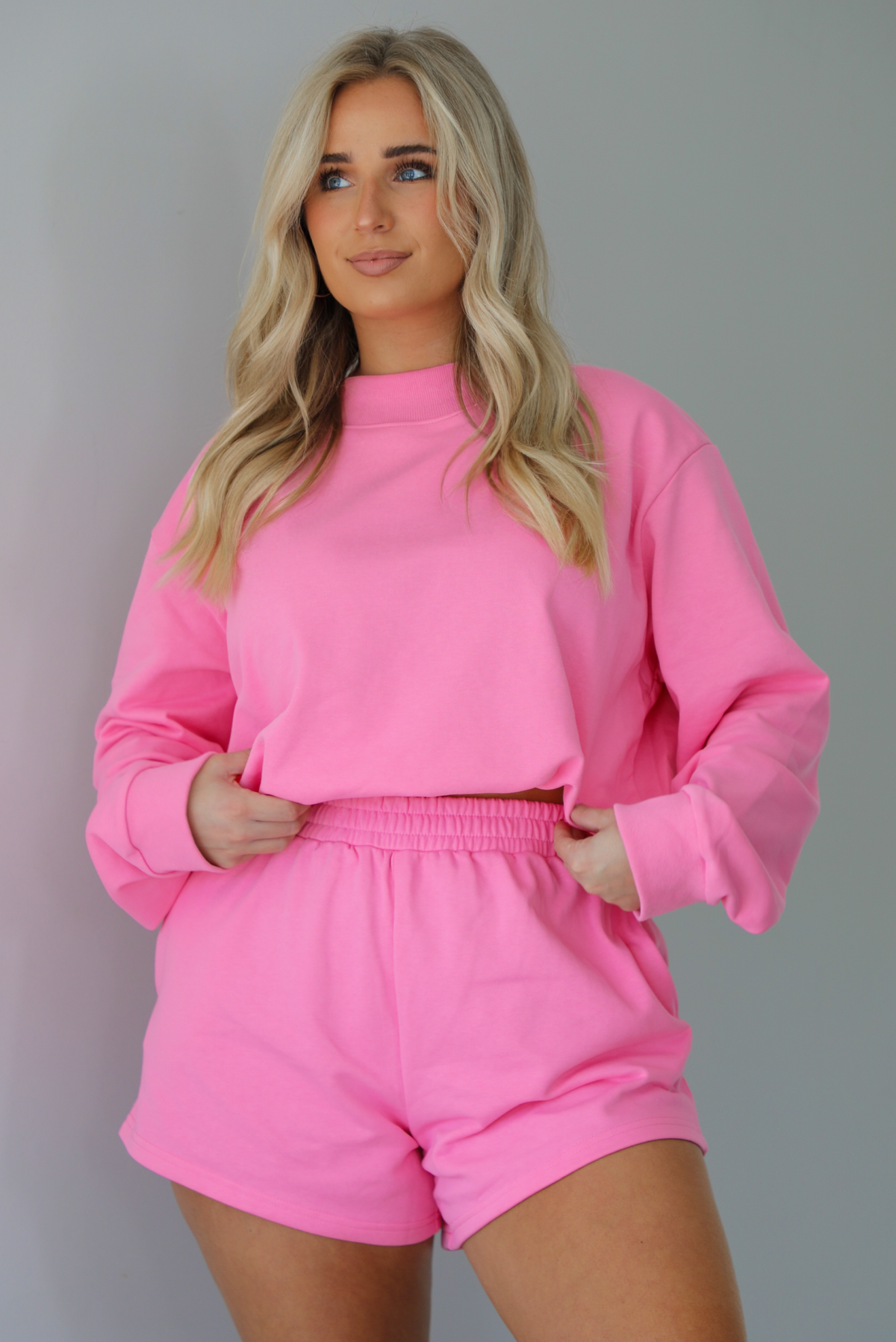 Woman wearing a hot pink two piece set featuring an oversized sweatshirt and high waisted shorts. The photo is taken indoors against a white wall and shows a closeup of the set.