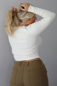 A woman shown from behind wearing a cream knit long sleeved top and high waisted olive-khaki cargo pants, standing indoors against a white wall.