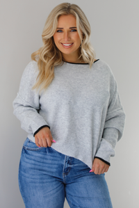 Woman wearing a grey relaxed fit sweater that has black trim on it, and medium wash distressed denim jeans. The photo is taken indoors against a white wall.
