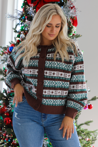 Close up of a woman model wearing a brown, green, and white Christmas themed sweater with multiple button closures in the front, slightly puffed sleeves, and a relaxed fit. The cardigan is paired with a white top underneath and medium wash denim jeans. The photo is taken inside a modern home with a decorated Christmas tree in the back. The photo shows the cardigan buttoned up.