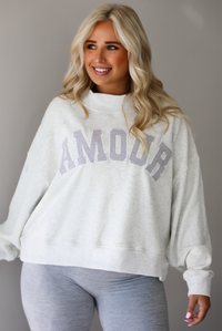 A woman posing in a light gray sweatshirt with 'AMOUR' printed in gray letters and light heather gray leggings, standing indoors against a white wall. The photo shows a closeup of the sweatshirt, showing how it has a high mock neck and is oversized. 
