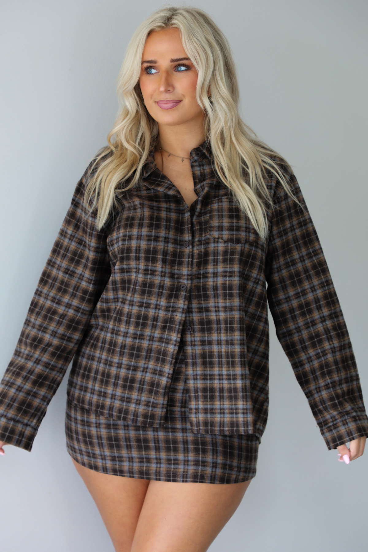 A woman wearing aa dark brown plaid set featuring an oversized button-down and fitted mini length skirt, standing indoors against a white wall.