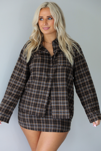 A woman wearing aa dark brown plaid set featuring an oversized button-down and fitted mini length skirt, standing indoors against a white wall.