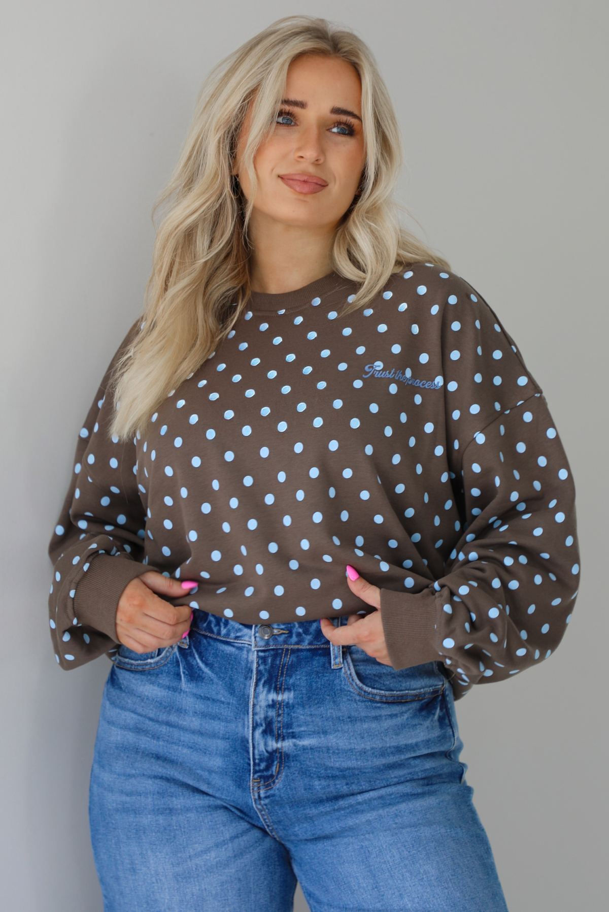 Woman wearing a brown sweater that has a blue polka dot print pattern throughout with blue embroidered writing on the left side that reads 'Trust The Process.' The sweater is paired with a medium wash denim. The photo shows a closeup of the sweater, which is folded under at the hem to give a cropped look. The photo is taken indoors against a white wall.