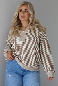 Woman wearing a mocha and cream striped pullover with a collared neckline, quarter button closures in the front, that has an oversized fit. The photo is taken indoors against a white wall.