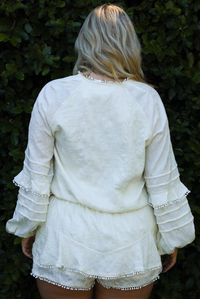 Back of a woman model wearing an off-white romper outdoors in front of greenery. 