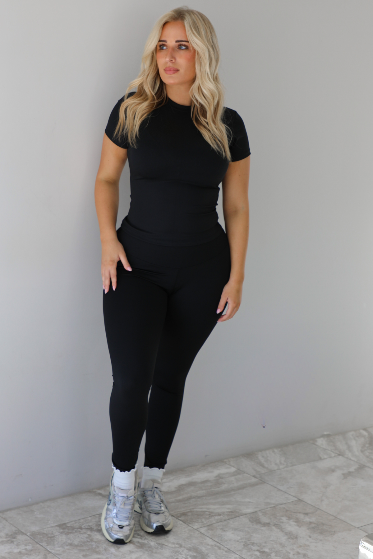 Woman wearing a fitted two piece black set featuring a short sleeved top and fitted leggings. The set is styled with a pair of white ankle socks and silver sneakers. The photo is taken indoors against a white wall with white marble flooring. 