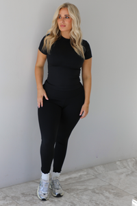 Woman wearing a fitted two piece black set featuring a short sleeved top and fitted leggings. The set is styled with a pair of white ankle socks and silver sneakers. The photo is taken indoors against a white wall with white marble flooring. 