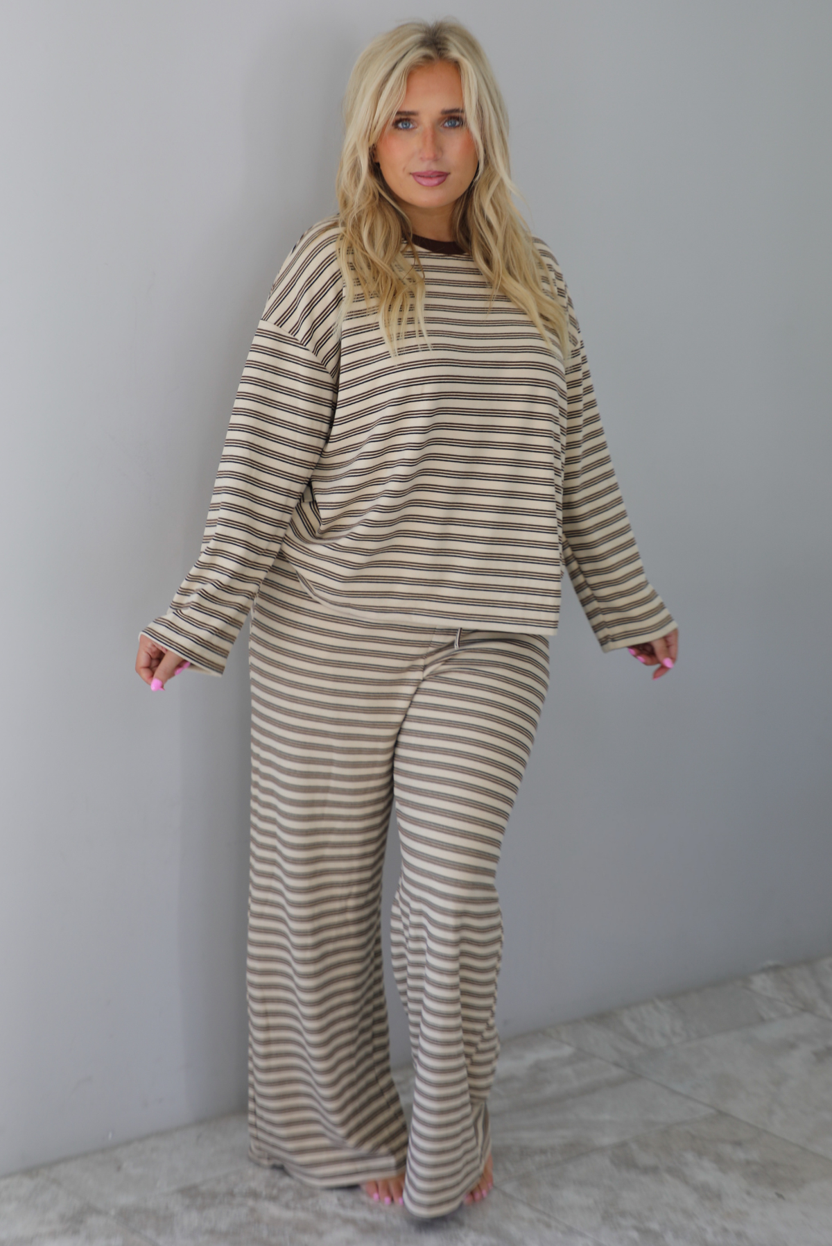 A woman wearing a sand colored matching two piece set with a brown striped pattern throughout featuring an oversized long sleeved top and oversized slouchy wide leg pants, standing indoors against a white wall with white marble flooring. 