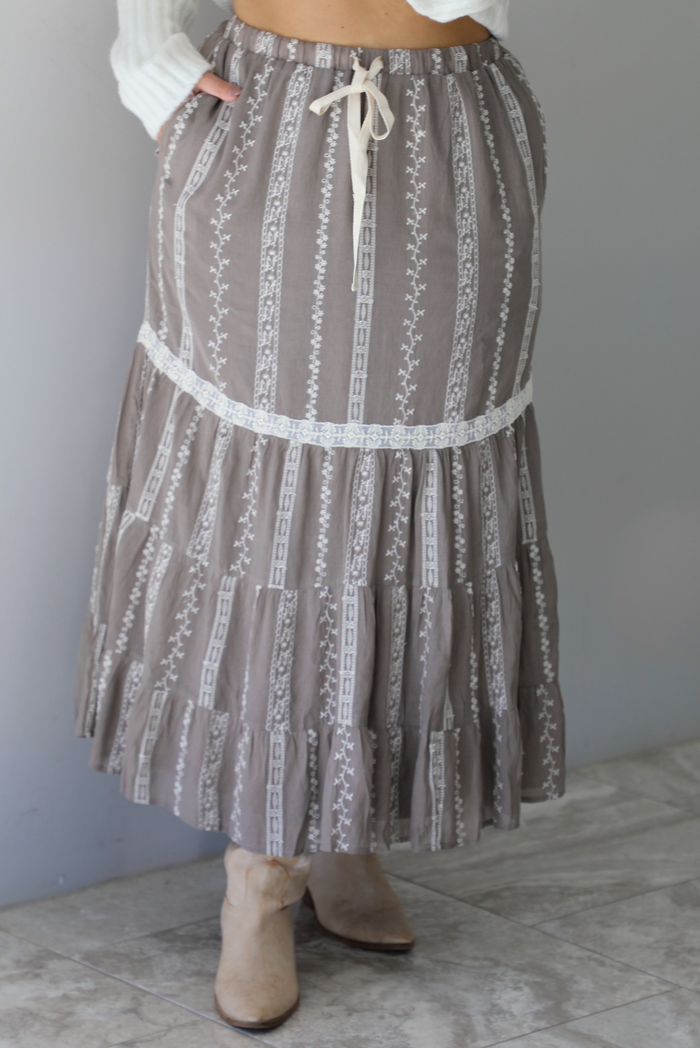 A closeup of a woman wearing a taupe with white floral embroidered pattern maxi skirt, standing indoors against a white wall with white marble flooring. 