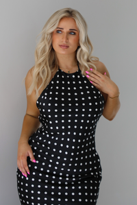 Woman wearing a black wearing a white polka dot printed midi dress that has a halter tie neckline, is form fitting, with white lace trim at the bottom. The photo is taken indoors against a white wall and shows a closeup of the bodice of the dress. 