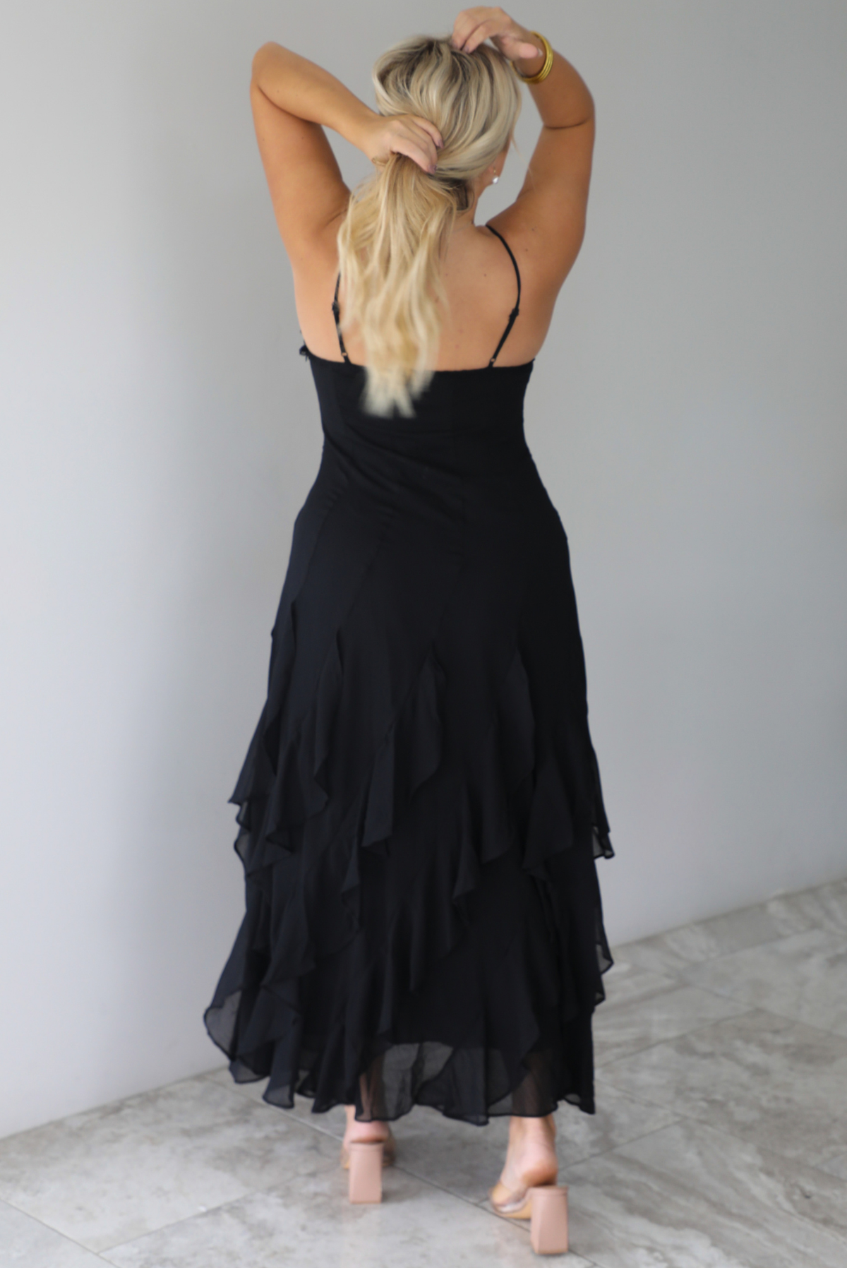 A woman shown from behind wearing a black formal maxi dress with lace trim detailing at the bodice and ruffled details at the skirt, standing indoors against a white wall with white marble flooring. The back shows the adjustable straps.