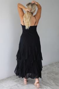 A woman shown from behind wearing a black formal maxi dress with lace trim detailing at the bodice and ruffled details at the skirt, standing indoors against a white wall with white marble flooring. The back shows the adjustable straps.