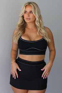 A blonde woman modeling a black two-piece activewear set consisting of a scoop-neck sports bra and a high-waisted mini skirt, both featuring thin white contrast piping details, indoors against a white wall.