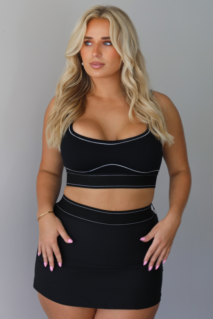 A blonde woman modeling a black two-piece activewear set consisting of a scoop-neck sports bra and a high-waisted mini skirt, both featuring thin white contrast piping details, indoors against a white wall.