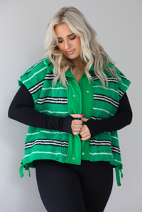 Woman wearing a green sherpa vest with black and white stripes that has an oversized fit, with a long sleeved, fitted, full body jumpsuit. The photo shows a close up of the vest with the functional button closures. The photo is taken indoors against a white wall with white marbled flooring. 