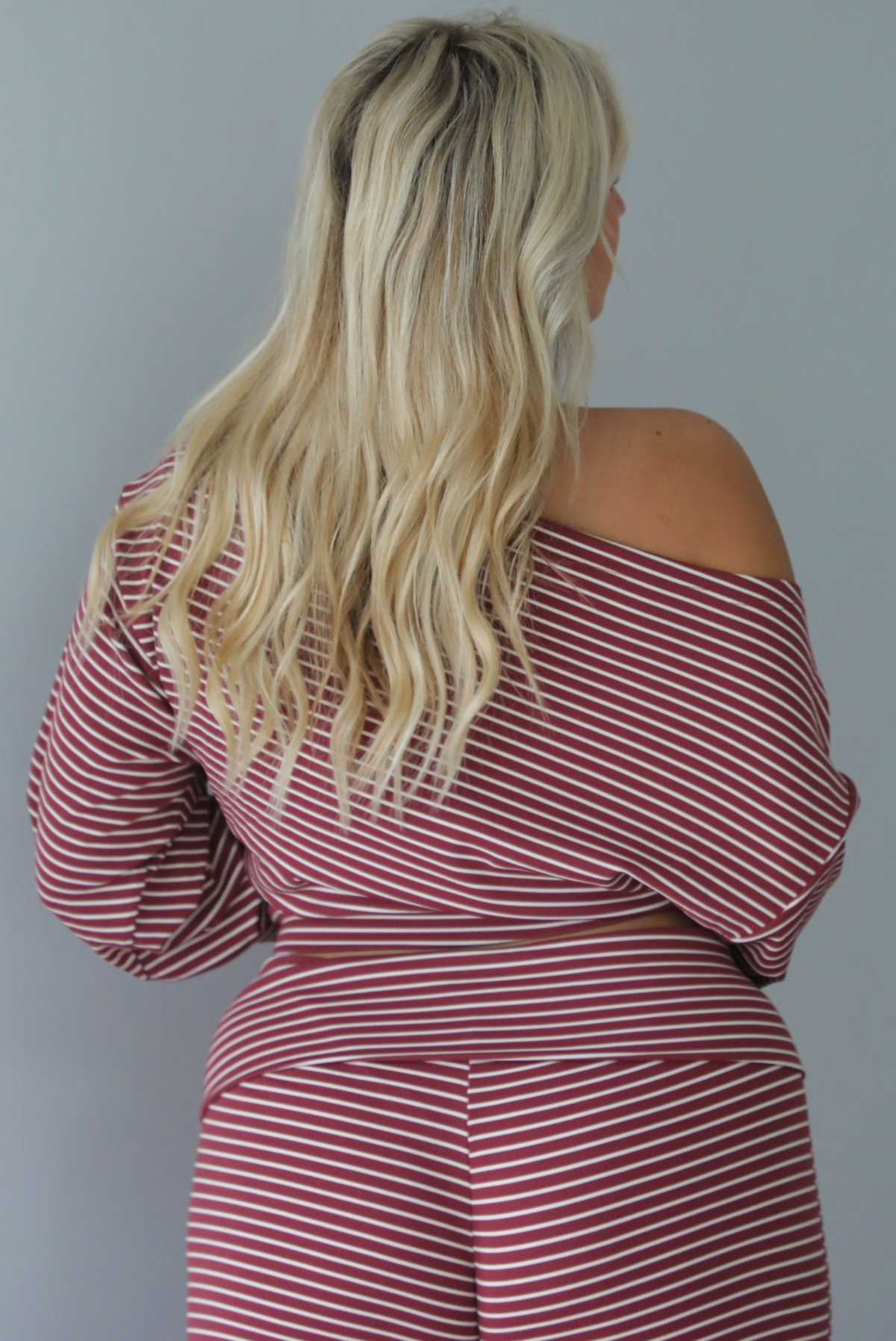 A woman shown from behind, focused on the back of the top, wearing a maroon and white striped two piece matching set featuring a slouchy off the shoulder long sleeved top and mid-rise, folded over waistline wide leg pants, standing indoors against a white wall.