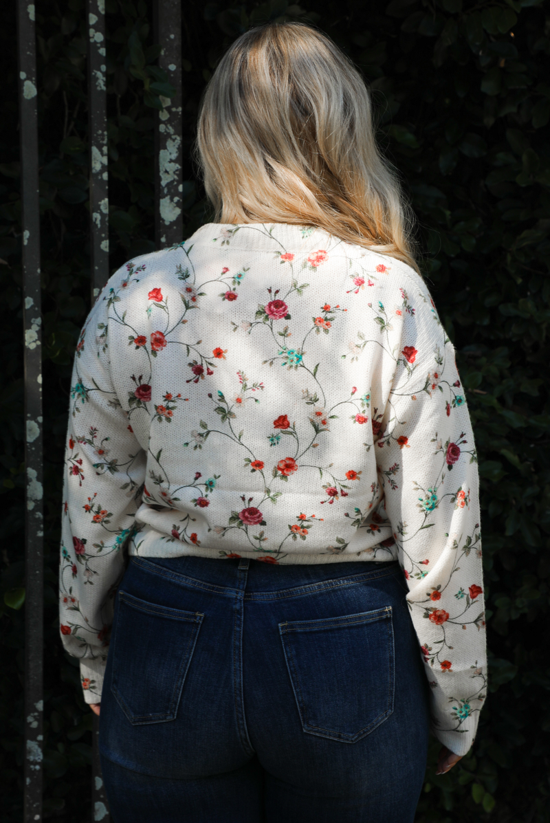 Woman model wearing an off-white sweater that has a multi-colored floral print pattern throughout with dark denim jeans. The photo showcases the length of the sweater, close up, in the back. The photo is taken next to an iron gate outdoors with greenery in the back. 