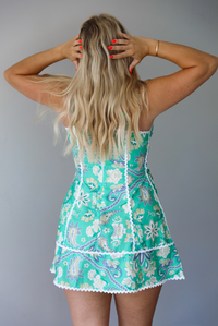 Woman wearing a teal green mini dress that has a multi-colored floral and paisley print pattern, white trim details, a plunging neckline, lace up details at the waist, and slightly flared skirt indoors against a white wall. The photo shows the back of the dress, which has a zipper closure. 