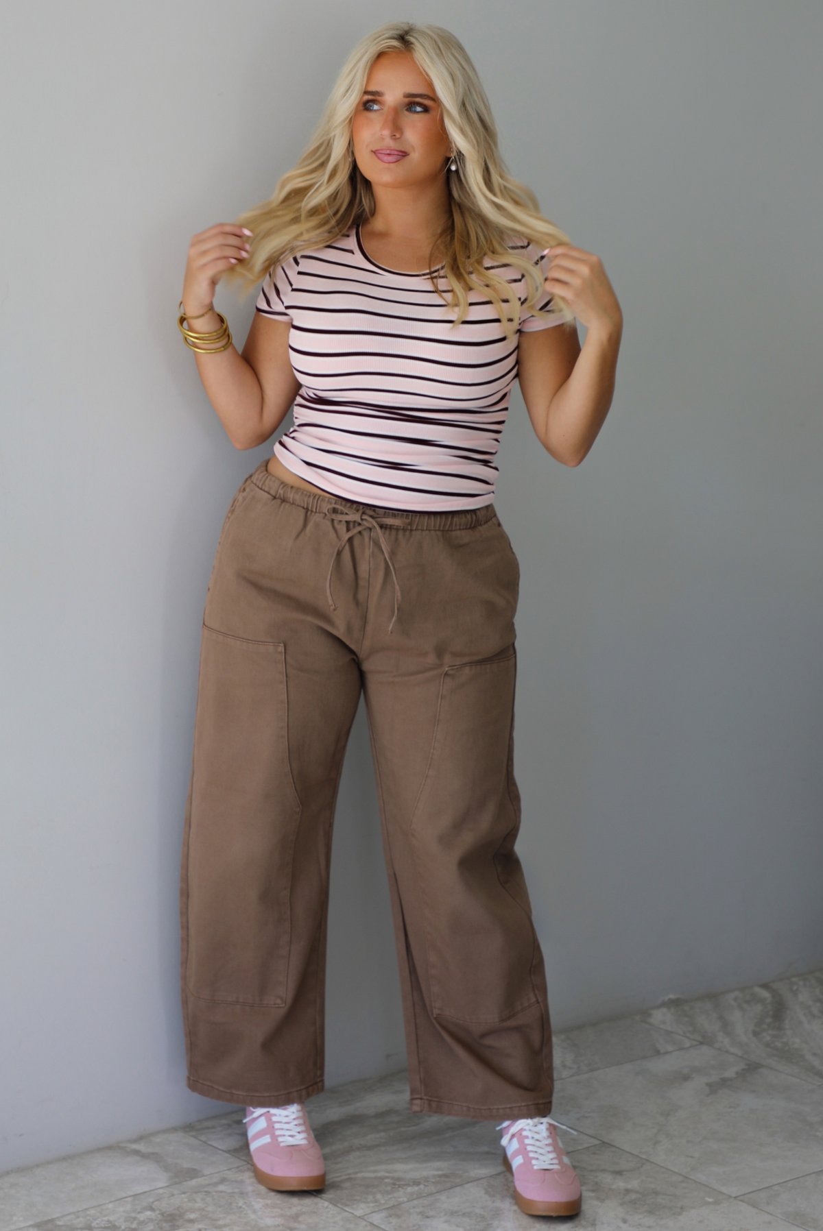 A woman wearing a pink baby tee that has a burgundy and white striped design throughout, paired with mocha barrel denim, standing indoors against a white wall with white marble flooring. 