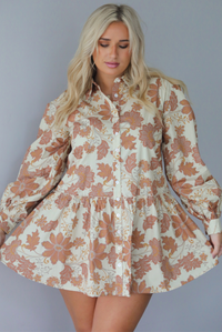 A woman wearing a cream dress with a tan fall floral print pattern throughout, collared neckline, functional button closures along the front, long sleeves that cinch tighter at the wrist, and a slightly flowy skirt, standing indoors against a white wall.
