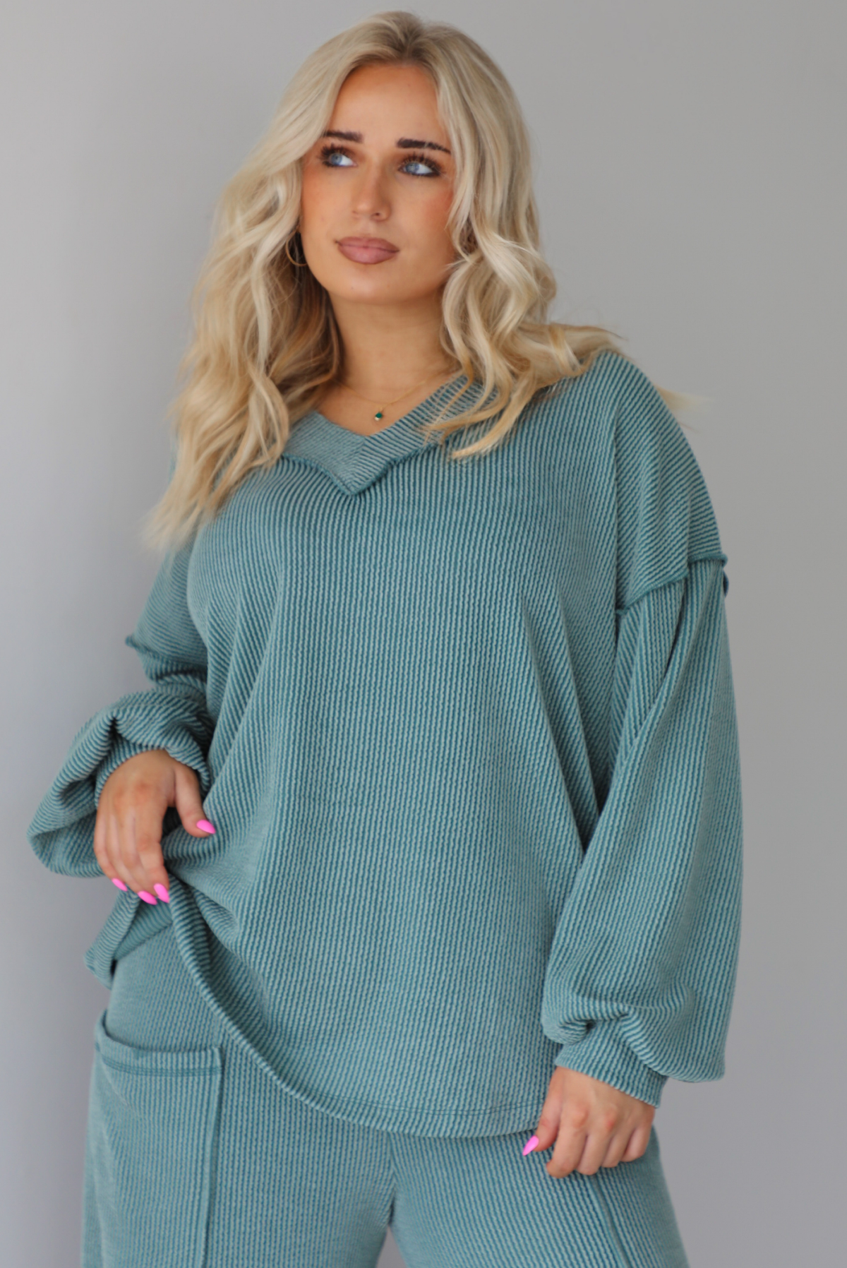 Woman wearing a teal two piece set that has a v-neck long sleeved oversized top and high waisted pants. The top is untucked to show its full length. The photo is taken indoors against a white wall.
