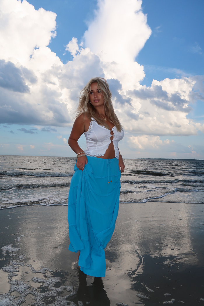 A woman stands on a beach wearing a white tank and a bright turquoise blue skirt, with ocean waves and a dramatic cloudy blue sky behind her.