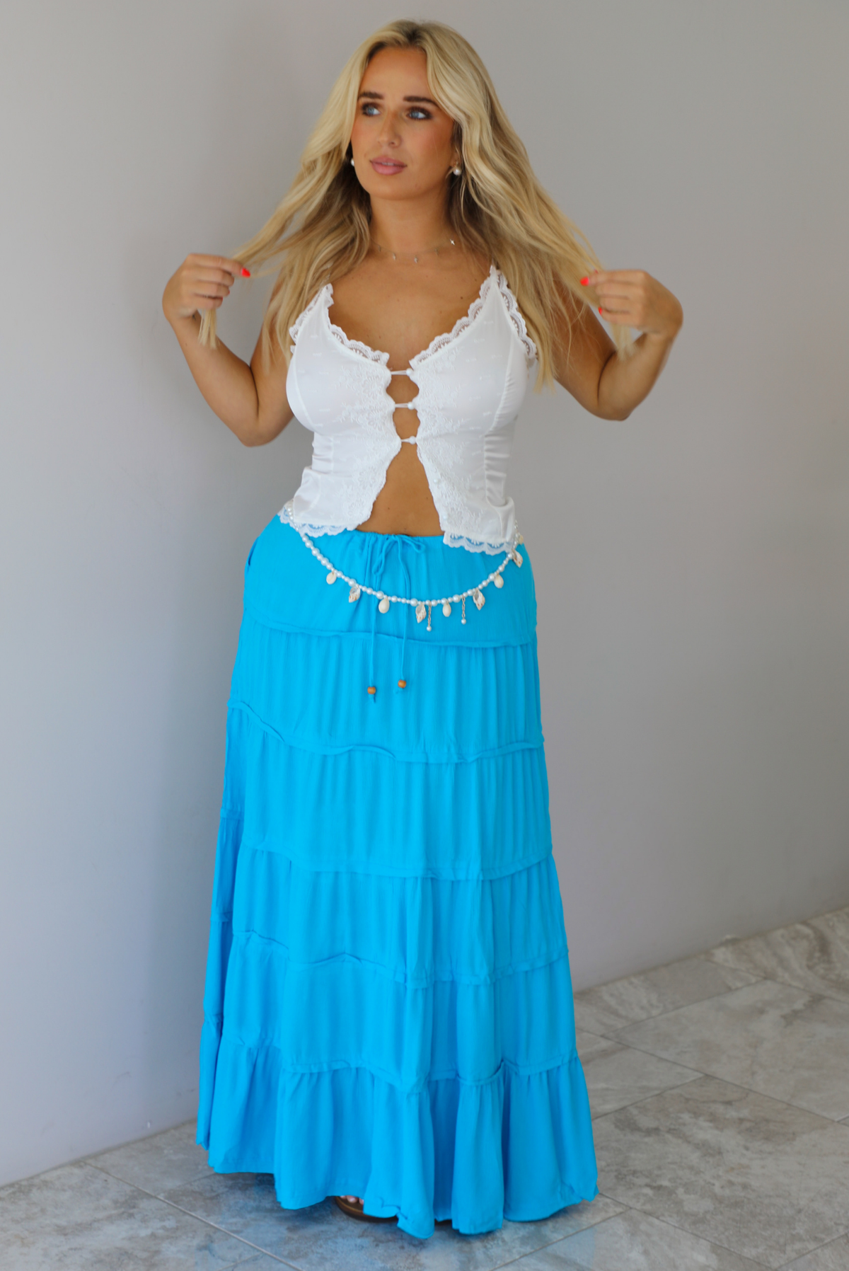A woman modeling a two-piece outfit consisting of a white lace-trimmed crop top and a bright turquoise blue tiered maxi skirt indoors against a white wall with white marble flooring. 