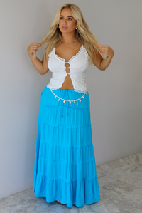 A woman modeling a two-piece outfit consisting of a white lace-trimmed crop top and a bright turquoise blue tiered maxi skirt indoors against a white wall with white marble flooring. 