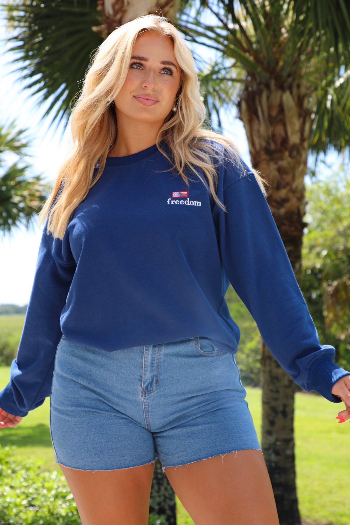 Woman wearing a navy sweatshirt that has FREEDOM embroidered on the front with medium wash denim shorts outdoors with greenery in the background.