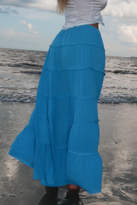 A tiered, long blue maxi skirt worn by a model standing on a beach with the ocean and cloudy sky in the background.