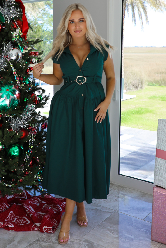 Woman model wearing a dark green midi length dress. It has a collared neckline, functional button closures in the front, and a belted detail at the waistline. The photo is taken inside a modern home with large windows and a decorated Christmas tree in the back. 