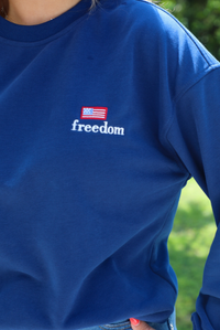 Woman wearing a navy sweatshirt that has FREEDOM embroidered on the front with medium wash denim shorts outdoors with greenery in the background. the photo shows a closeup of the embroidered detail in the front that reads 'freedom' and has an American Flag stitched above. 