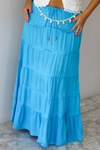 A tiered maxi skirt in bright turquoise blue, featuring a drawstring waist, worn by a model indoors against a white wall with white marble flooring. The model has paired the skirt with a pearl and shell belly chain/belt. 