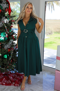 Woman model wearing a dark green midi length dress. It has a collared neckline, functional button closures in the front, and a belted detail at the waistline. The photo is taken inside a modern home with large windows and a decorated Christmas tree in the back. 
