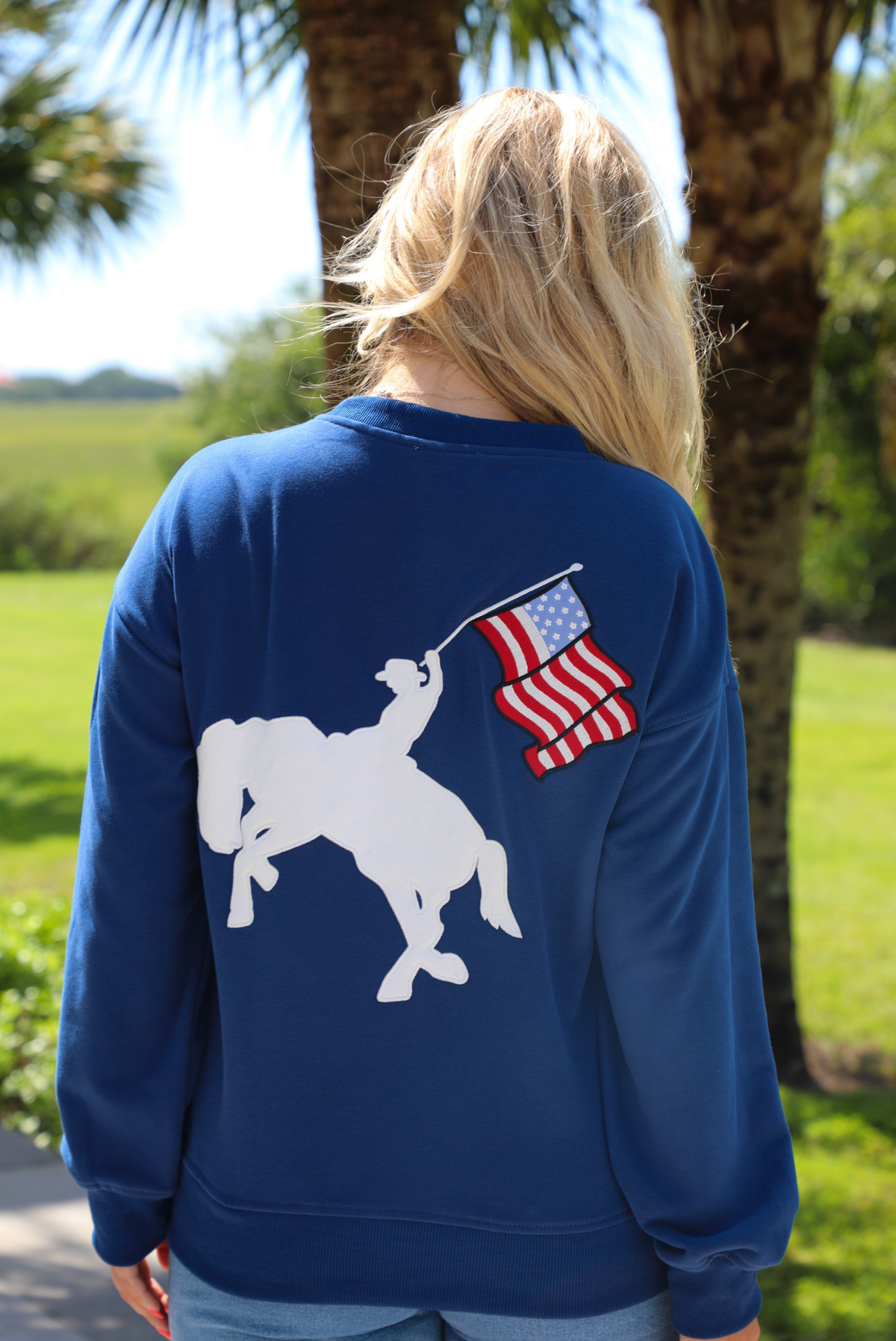 Woman wearing a navy sweatshirt that has FREEDOM embroidered on the front with medium wash denim shorts outdoors with greenery in the background. The photo shows a closeup of the back, which depicts an embroidered design of a  person riding a horse while holding an American Flag. 