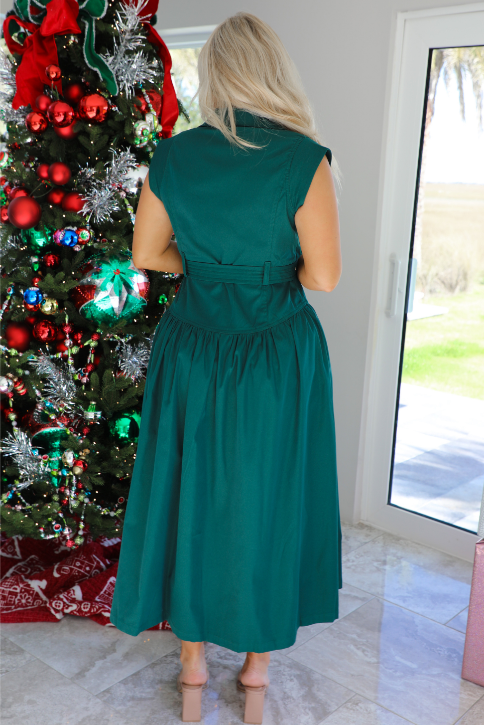 Back of a woman model wearing a dark green midi length dress. It has a collared neckline, functional button closures in the front, and a belted detail at the waistline. The photo is taken inside a modern home with large windows and a decorated Christmas tree in the back. 
