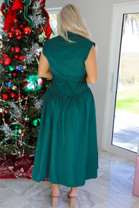 Back of a woman model wearing a dark green midi length dress. It has a collared neckline, functional button closures in the front, and a belted detail at the waistline. The photo is taken inside a modern home with large windows and a decorated Christmas tree in the back. 