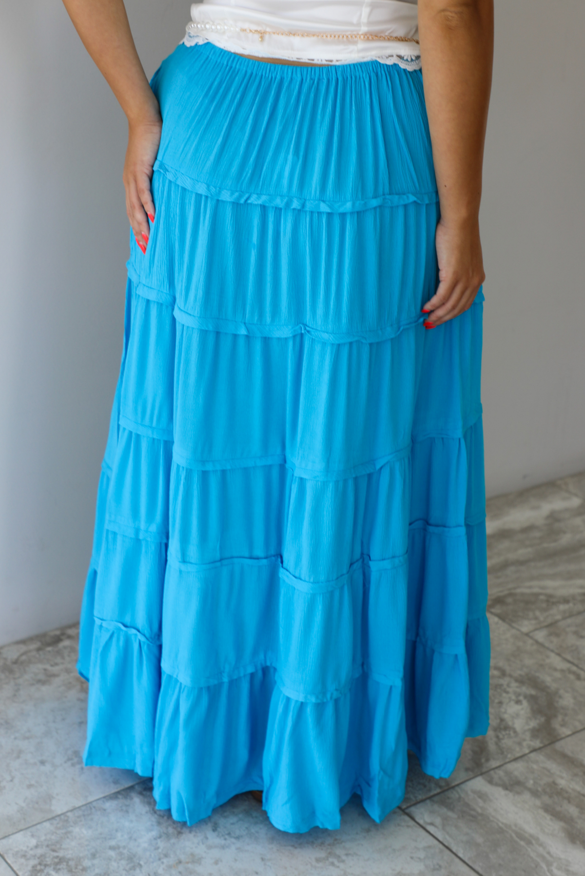 A tiered maxi skirt in bright turquoise blue, featuring a drawstring waist, worn by a model indoors against a white wall with white marble flooring. The photo shows the back of the skirt. 
