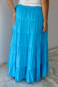 A tiered maxi skirt in bright turquoise blue, featuring a drawstring waist, worn by a model indoors against a white wall with white marble flooring. The photo shows the back of the skirt. 