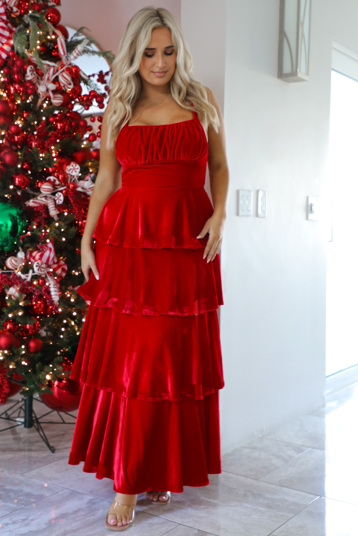Woman model wearing a red velvet textured ruffled maxi dress. The dress has adjustable straps, a fitted waistline, and a ruffled skirt. The photo is taken inside a modern home with a decorated Christmas tree in the back. 