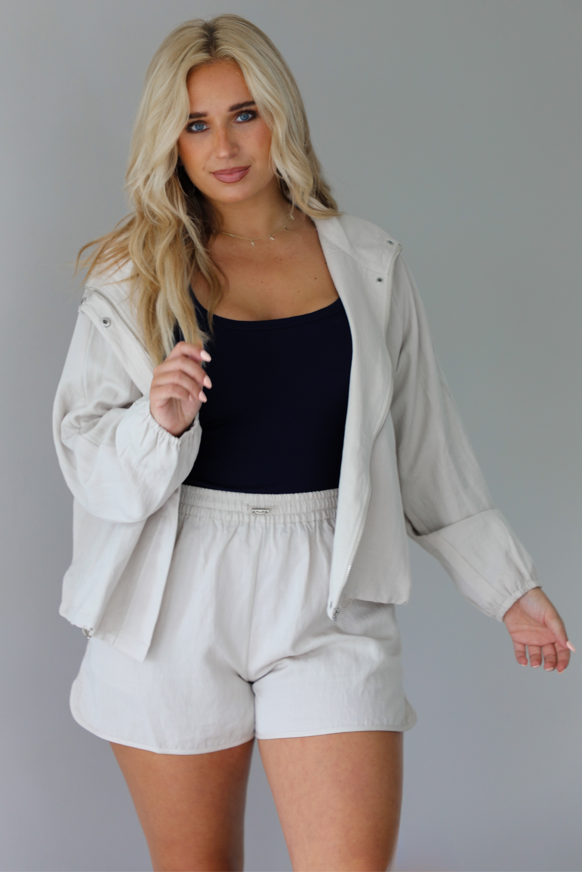 Woman wearing a stone colored two piece set featuring a zip up jacket and shorts with a navy tank tucked into the shorts indoors against a white wall.