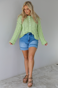 Woman wearing a green, long sleeved sweater with a loose knit design, functional button closures in the front, cuffed sleeves, and a raw, unfinished hem with medium wash denim shorts indoors against a white wall with white marble flooring. 
